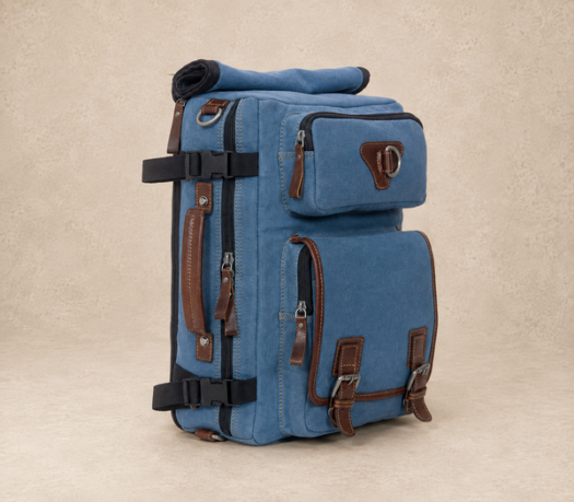 Blue canvas travel backpack with brown leather trim and black webbing, AIL-BP-0025, shown from a front three-quarter angle on a warm limestone studio background