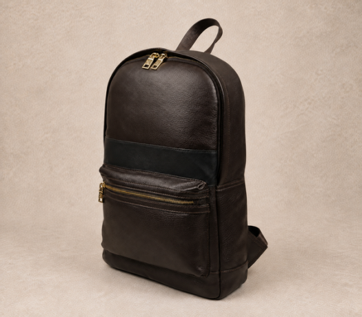 Dark brown pebbled leather backpack with black trim, AIL-BP-0026, shown from a front three-quarter angle on a warm limestone studio background