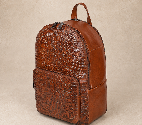 Cognac brown leather backpack with crocodile-embossed front panels, AIL-BP-0028, shown from a front three-quarter angle on a warm limestone studio background