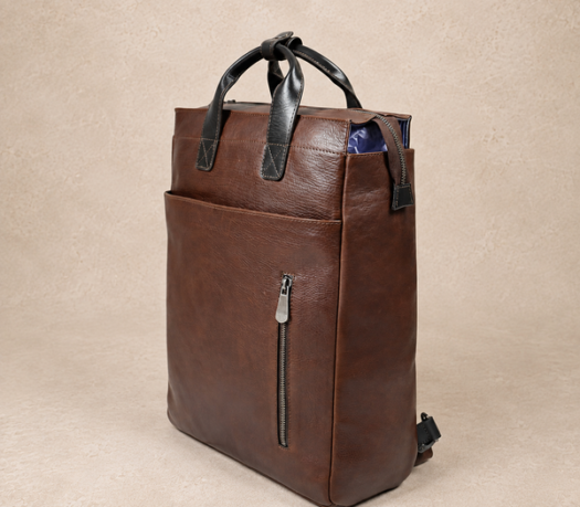 Brown leather backpack with black carry handles, AIL-BP-0067, shown from a front three-quarter angle on a warm limestone studio background