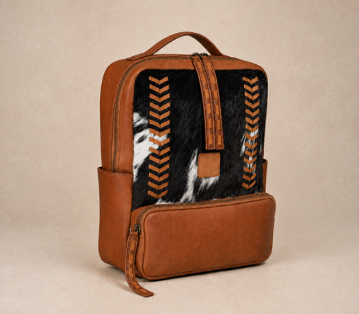 Cognac brown leather backpack with a black-and-white hair-on-hide front panel, AIL-BP-0086, shown from a front three-quarter angle on a warm limestone studio background