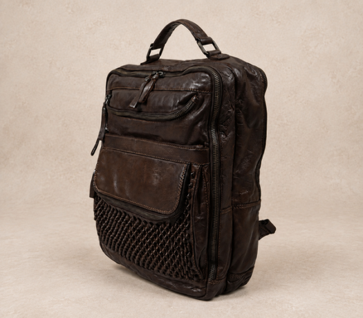 Dark brown distressed leather backpack with a woven leather front panel, AIL-BP-0090, shown from a front three-quarter angle on a warm limestone studio background